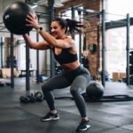 Na tym obrazie znajduje się wysportowana kobieta z dynamicznie związanymi w kucyk włosami, ubrana w czarny sportowy top oraz szare legginsy